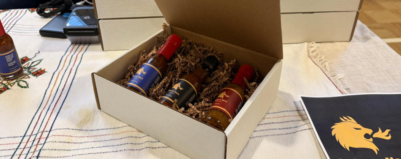 An open cardboard box containing three bottles of hot sauce, resting on crinkle paper, with other boxes and a payment processing device on a Ethiopian style tablecloth.